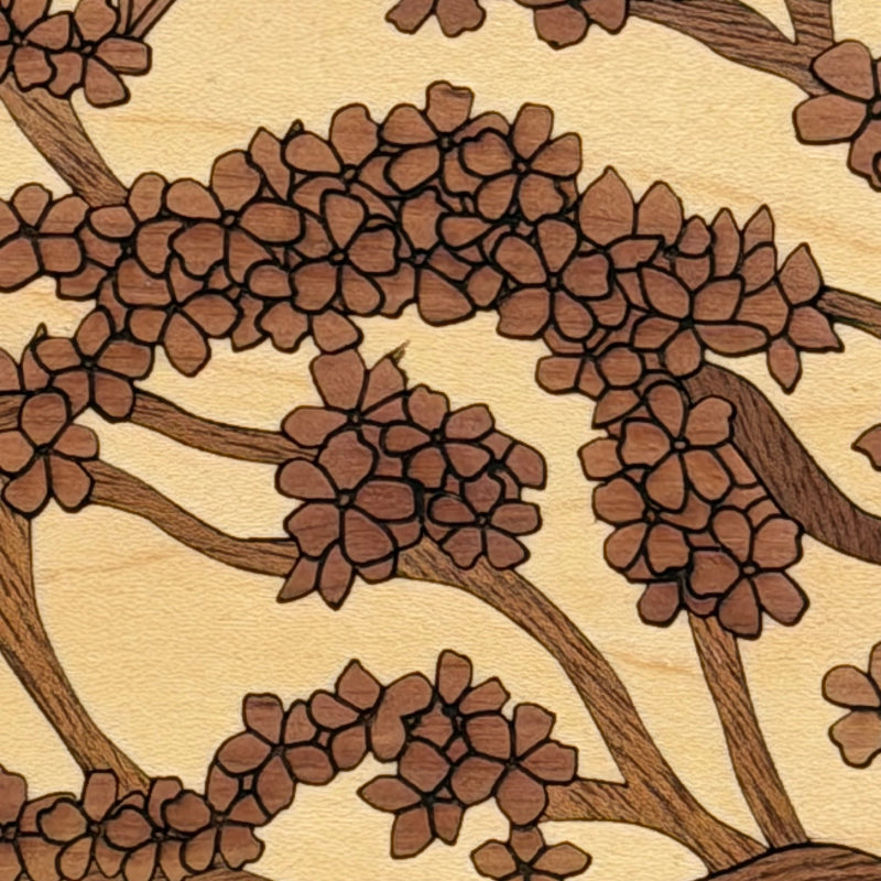 A close-up of a wood grain surface featuring an engraved or burned floral pattern with brown blossoms and branches on a light beige background.