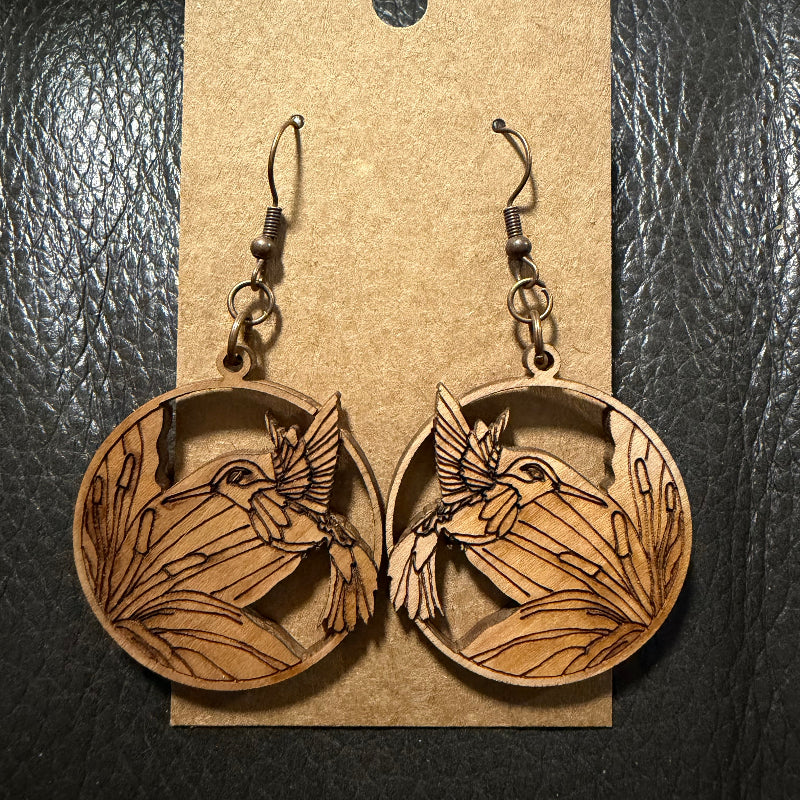 A pair of oval wooden dangle earrings with laser-engraved hummingbird and reeds designs, displayed on a kraft paper hanger against a black textured background.
