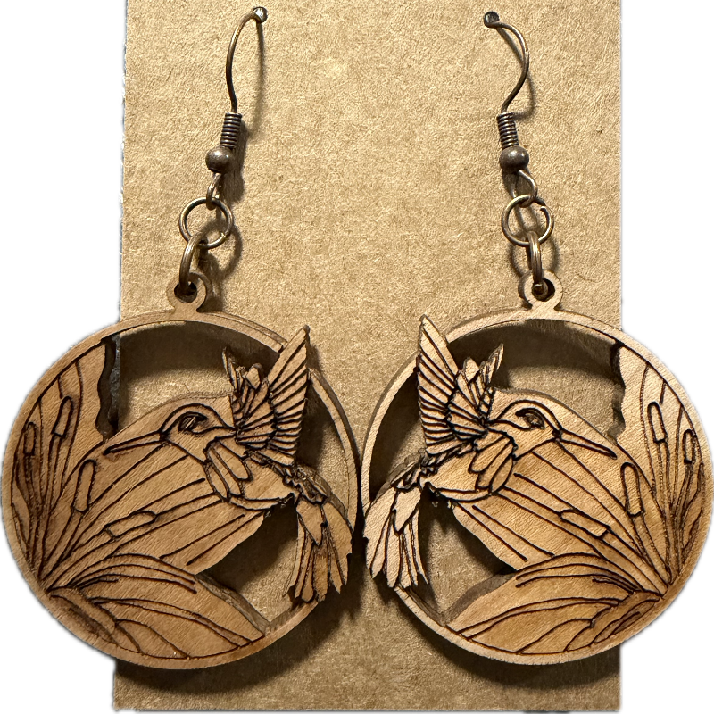 A pair of oval wooden dangle earrings with engraved bird and leaf designs, hanging from dark metal hooks on a kraft paper display card.