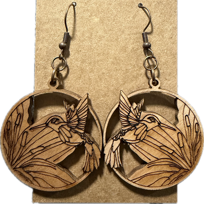 A pair of oval wooden dangle earrings with engraved bird and leaf designs, hanging from dark metal hooks on a kraft paper display card.