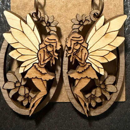 A pair of laser-cut wooden fairy earrings displayed on a brown kraft card against a dark slate background.