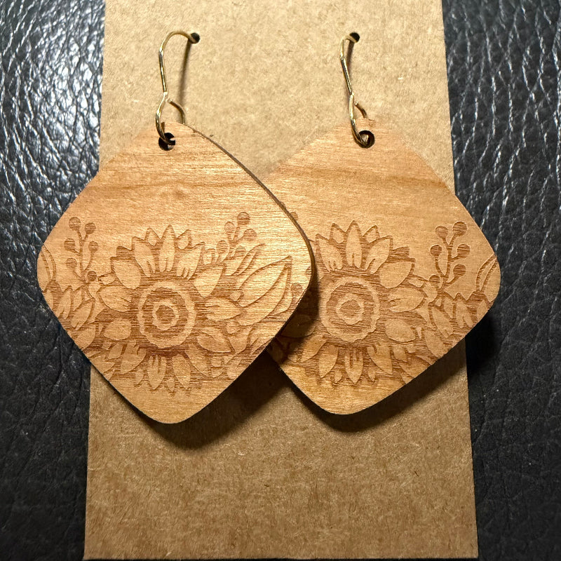 A pair of wooden dangle earrings with a carved floral sunflower design, displayed on a brown kraft paper card against a black textured background.