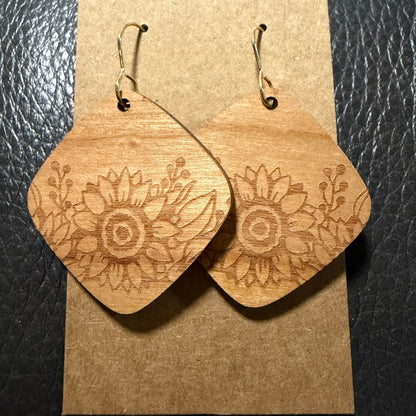 A pair of wooden dangle earrings with a carved floral sunflower design, displayed on a brown kraft paper card against a black textured background.