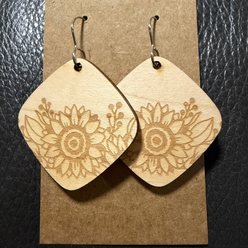 A pair of wooden dangle earrings with a sunflower engraving, displayed on a brown kraft paper card against a black textured background.