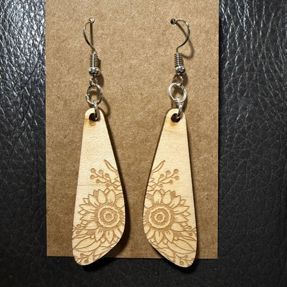 A pair of teardrop-shaped wooden dangle earrings with laser-engraved floral sunflower patterns, hanging from silver-tone fishhook wires on a brown kraft paper display card against a black textured background.
