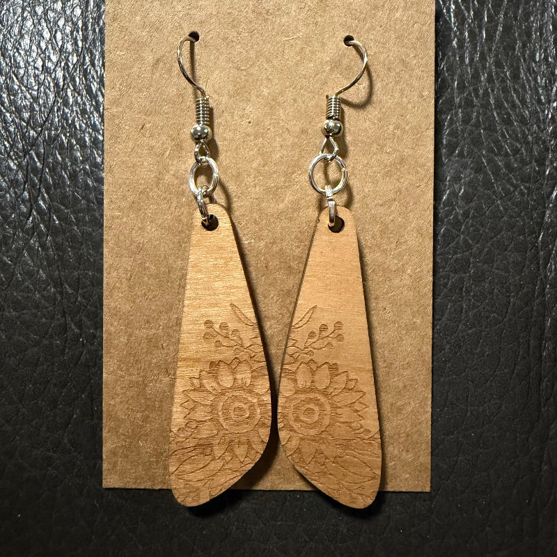 A pair of wooden dangle earrings with engraved floral patterns, hanging from silver fishhook wires on a brown kraft paper display card against a black textured background.