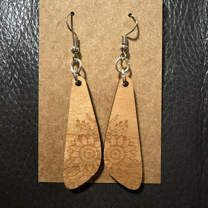 A pair of wooden dangle earrings with engraved floral patterns, hanging from silver fishhook wires on a brown kraft paper display card against a black textured background.