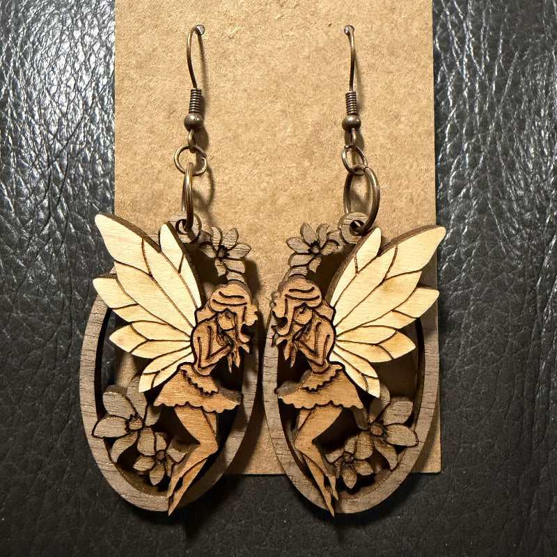 A pair of wooden fairy drop earrings with laser-cut details, hanging from dark metal fishhook wires against a brown kraft paper display card, set on a dark textured background.