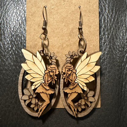 A pair of wooden fairy drop earrings with laser-cut details, hanging from dark metal fishhook wires against a brown kraft paper display card, set on a dark textured background.