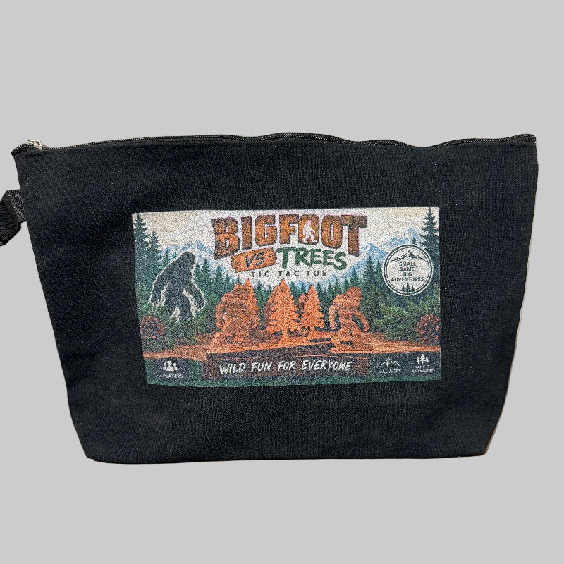 A black canvas zippered pouch with a colorful 'Bigfoot vs Trees' graphic on the front, sitting on a wooden table against a light textured wall.