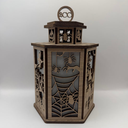 A hexagonal wooden lantern with Halloween-themed laser-cut details including spiders, cobwebs, and jack-o'-lantern windows, topped with a circular 'BOO' cutout.