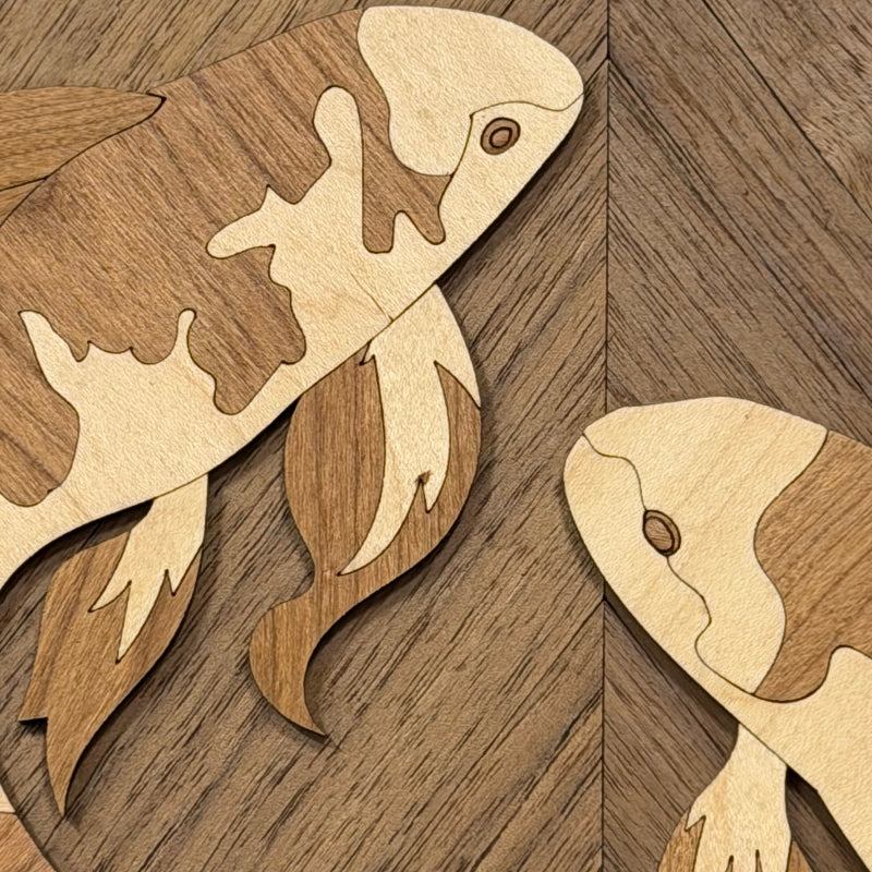 Close-up of wooden fish-shaped pieces with a light natural wood body and darker brown etched or inlaid patterns, arranged on a wood-grain surface.