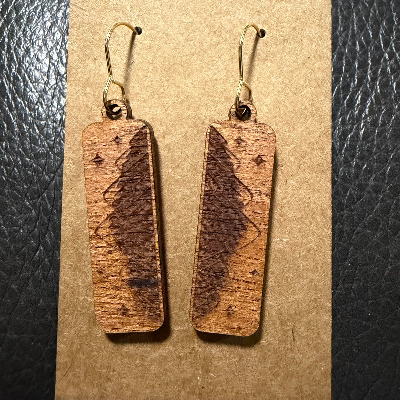 A pair of rectangular wooden drop earrings with a dark engraved tree design and small star motifs, hanging from gold-tone fishhook wires on a brown kraft card against a black textured background.