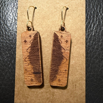 A pair of rectangular wooden drop earrings with a dark engraved tree design and small star motifs, hanging from gold-tone fishhook wires on a brown kraft card against a black textured background.