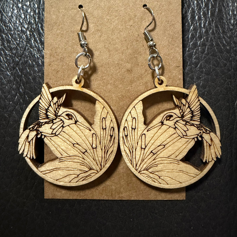A pair of wooden dangle earrings with a laser-cut hummingbird and reeds design, hanging from silver fishhook wires on a brown kraft paper display card, set against a black textured background.
