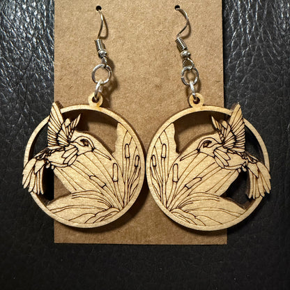A pair of wooden dangle earrings with a laser-cut hummingbird and reeds design, hanging from silver fishhook wires on a brown kraft paper display card, set against a black textured background.