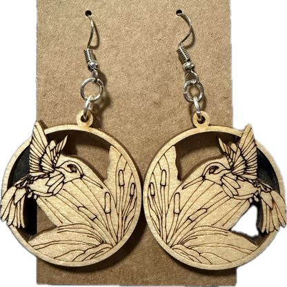 A pair of wooden dangle earrings displayed on a brown kraft card. Each circular wooden earring features a laser-cut design of a bird in flight above reeds, with a silver-toned fishhook ear wire attached at the top.