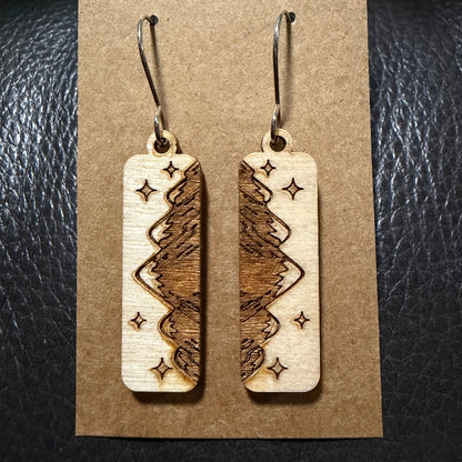 A pair of rectangular wooden dangle earrings with a laser-engraved tree and star pattern, displayed on a brown kraft paper card against a black textured leather background.
