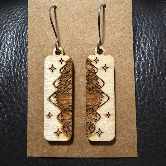 A pair of rectangular wooden dangle earrings with a laser-engraved tree and star pattern, displayed on a brown kraft paper card against a black textured leather background.