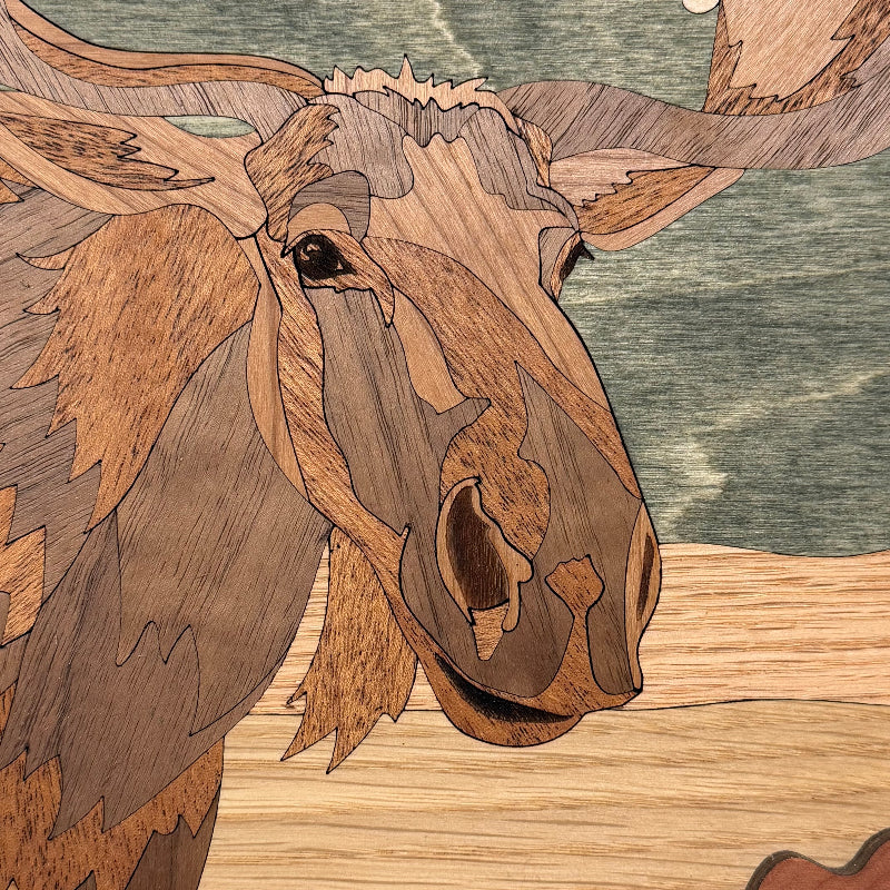 Close-up of a moose head artwork made from layered wood veneers, showing natural wood grain patterns in shades of brown and tan against a muted blue-green background.
