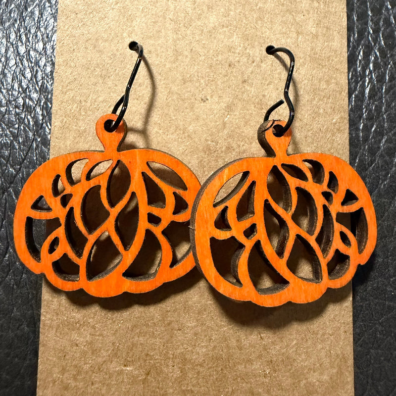 A pair of orange laser-cut wooden pumpkin earrings with black wire hooks, displayed on a brown kraft paper background.