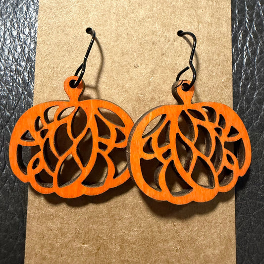 A pair of orange laser-cut wooden pumpkin earrings with black wire hooks, displayed on a brown kraft paper background.