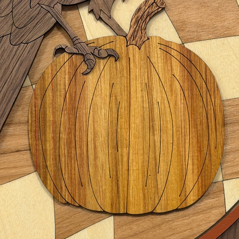 A close-up of a wooden pumpkin-shaped decorative piece with engraved wood grain and black line details, placed on a multicolor wood-patterned surface.