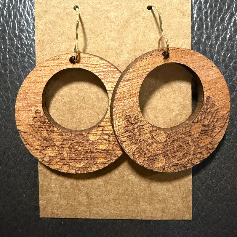 A pair of large circular wooden drop earrings with floral laser-engraved patterns and gold-tone fishhook wires, displayed on a brown kraft paper card against a black textured background.