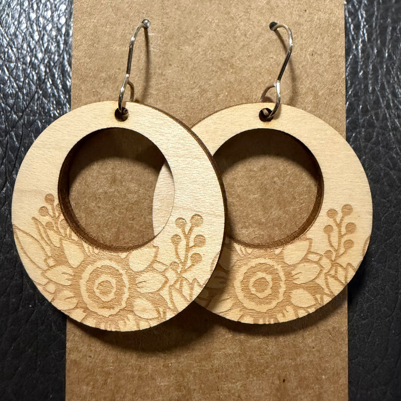 A pair of large circular wooden dangle earrings with laser-engraved floral patterns, hanging from silver fishhook wires on a brown kraft paper backing card.