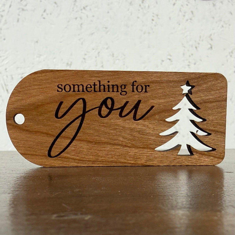 A wooden ornament-shaped sign with a hole on the left, featuring the engraved text 'something for you' in black script and a white laser-cut Christmas tree with a star on top, displayed on a wooden surface against a textured white wall.
