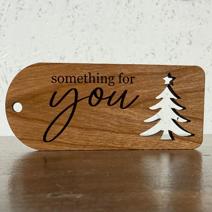 A wooden ornament-shaped sign with a hole on the left, featuring the engraved text 'something for you' in black script and a white laser-cut Christmas tree with a star on top, displayed on a wooden surface against a textured white wall.