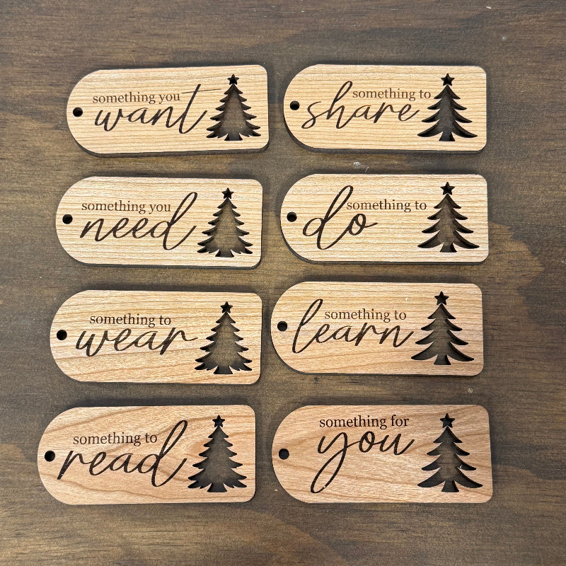 Eight small rectangular wooden tags with rounded corners are arranged in a 2-by-4 grid on a dark wood surface. Each tag has engraved black text reading a different phrase, such as "something you want," "something to share," "something you need," "something to do," "something to wear," "something to learn," "something to read," "something for you," plus a black tree cut out accent.