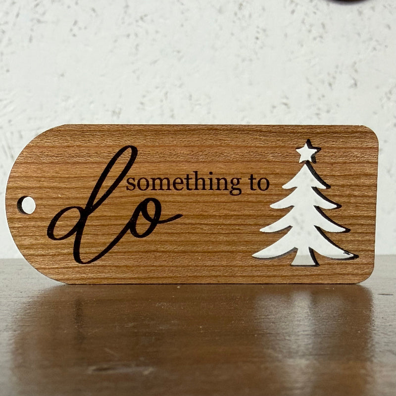 A wooden plaque with a rounded rectangular shape, featuring the engraved text 'something to do' in black, alongside a white silhouette of a Christmas tree with a star on top. The plaque is resting on a dark wooden surface against a textured off-white wall.