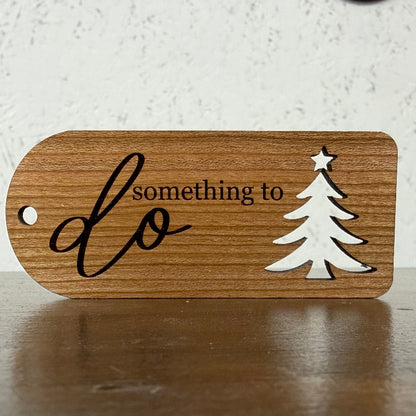 A wooden plaque with a rounded rectangular shape, featuring the engraved text 'something to do' in black, alongside a white silhouette of a Christmas tree with a star on top. The plaque is resting on a dark wooden surface against a textured off-white wall.