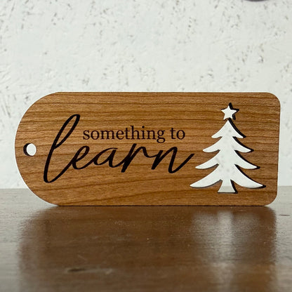 A rectangular wooden tag with rounded corners, featuring the engraved text 'something to learn' in black script and a white silhouette of a Christmas tree with a star. The tag is shown resting on a dark wooden surface against a light, textured wall.