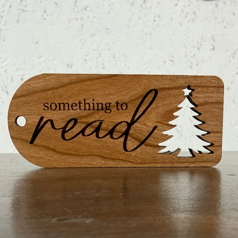 A wooden bookish tag with a rounded shape, featuring the engraved text 'something to read' and a white silhouette of a Christmas tree, resting on a wooden surface against a textured white wall.