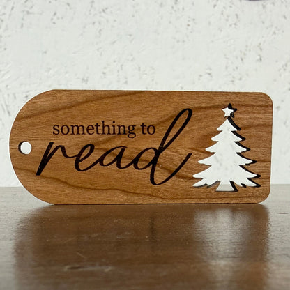 A wooden bookish tag with a rounded shape, featuring the engraved text 'something to read' and a white silhouette of a Christmas tree, resting on a wooden surface against a textured white wall.