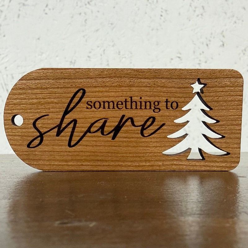A wooden tag with rounded corners, featuring the engraved text 'something to share' in black script and a white silhouette of a Christmas tree with a star, resting on a dark wooden surface.