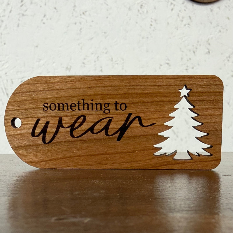 A wooden ornament or gift tag with a rounded rectangular shape, featuring the engraved text 'something to wear' in black, a white silhouette of a Christmas tree, and a hole on the left side, displayed on a wooden surface.