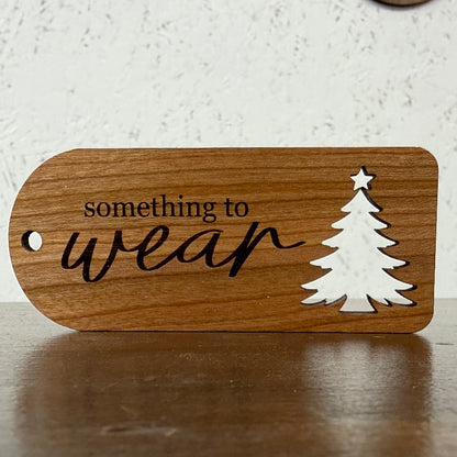 A wooden ornament or gift tag with a rounded rectangular shape, featuring the engraved text 'something to wear' in black, a white silhouette of a Christmas tree, and a hole on the left side, displayed on a wooden surface.