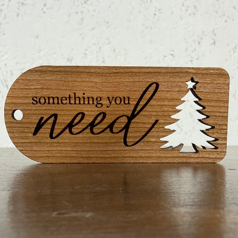A wooden gift tag with rounded corners, featuring the engraved text 'something you need' in black script and a white silhouette of a Christmas tree with a star on top. The tag is displayed on a dark wooden surface against a light, textured background.