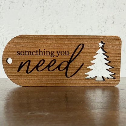 A wooden gift tag with rounded corners, featuring the engraved text 'something you need' in black script and a white silhouette of a Christmas tree with a star on top. The tag is displayed on a dark wooden surface against a light, textured background.