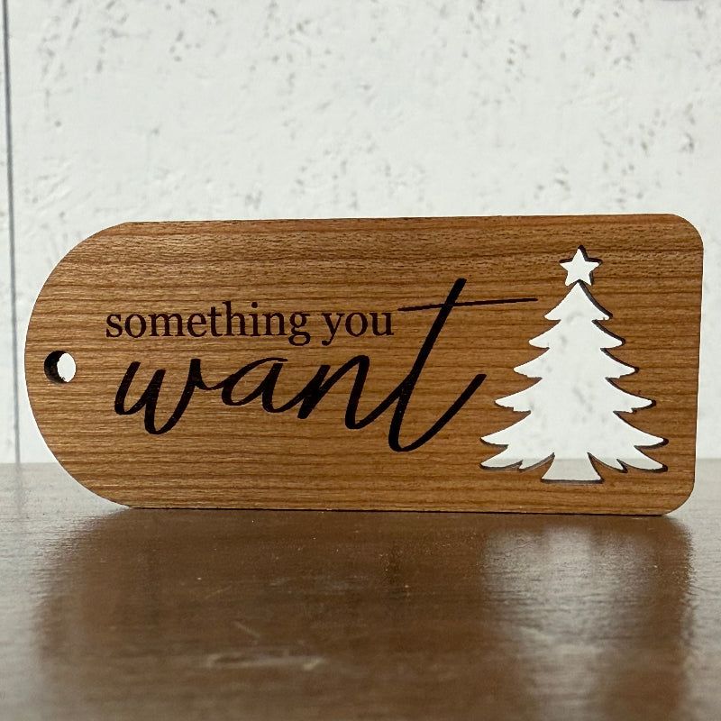 A wooden ornament tag with a rounded rectangular shape, featuring the engraved text 'something you want' in black, alongside a white silhouette of a Christmas tree with a star on top. The tag is displayed on a dark wooden surface against a light, textured background.