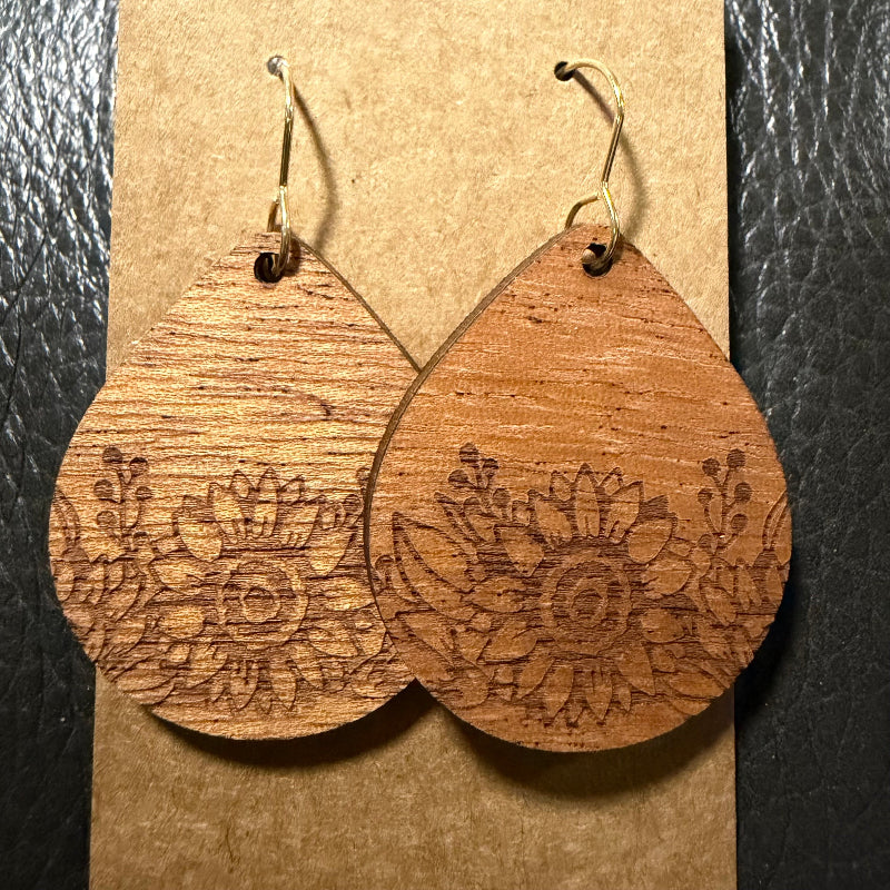 A pair of teardrop-shaped wooden dangle earrings with engraved floral patterns and gold-tone hooks, displayed on a brown kraft card against a black textured background.