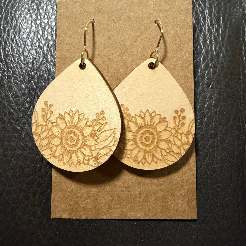A pair of teardrop-shaped wooden dangle earrings with engraved floral sunflower patterns, displayed on a brown kraft paper card against a black textured background.