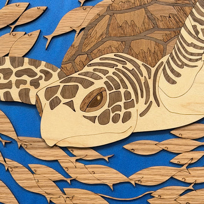 A close-up of a wooden turtle and fish cutout artwork, showing layered laser-cut wood pieces in natural tan tones over a bright blue background.